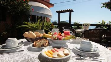 A table set for breakfast with various foods, including bread, fruits, and eggs, on a terrace with a view of the sea and mountains.