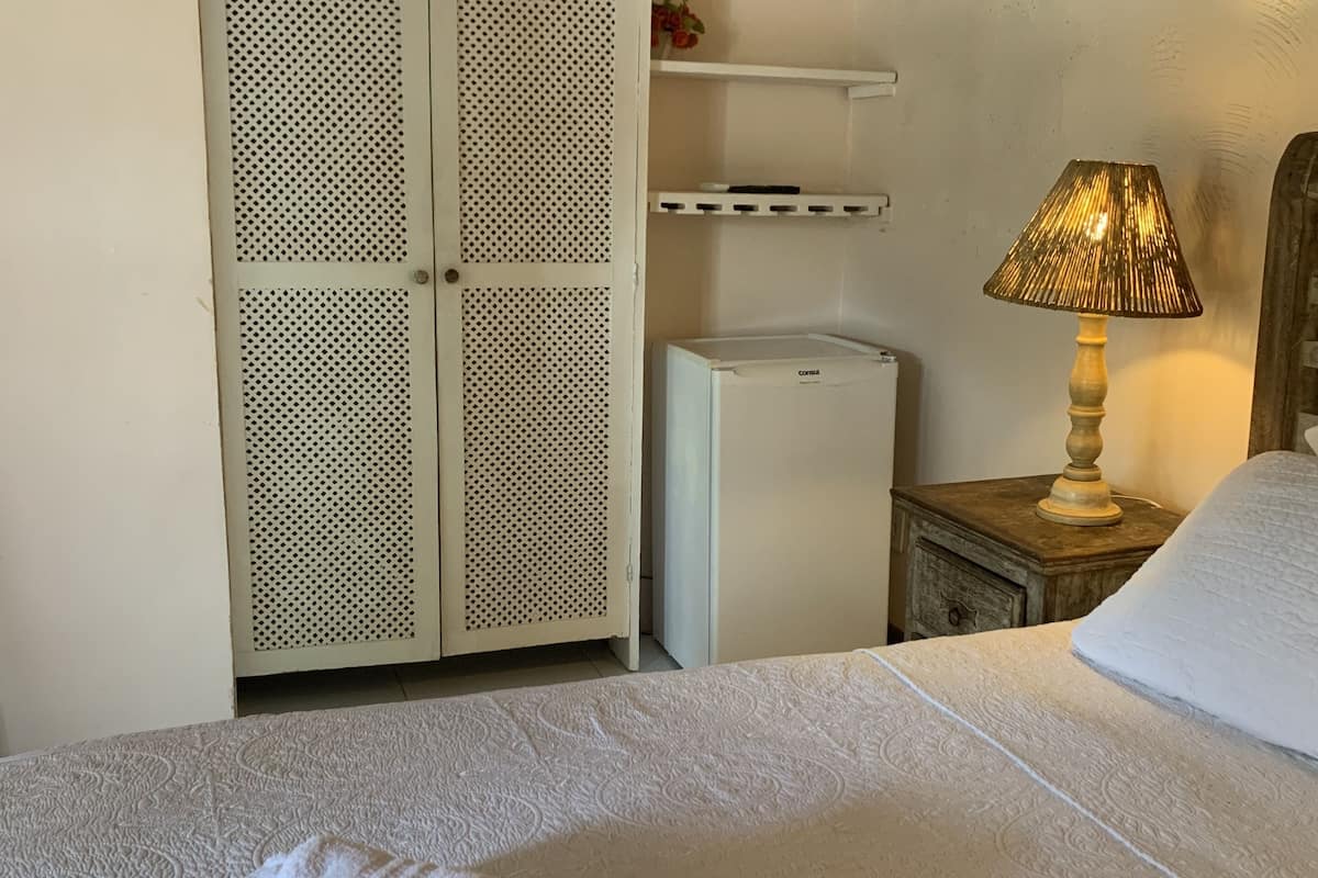 A hotel room with a bed, a wardrobe, a small refrigerator, a bedside table with a lamp, and a bedside pillow.