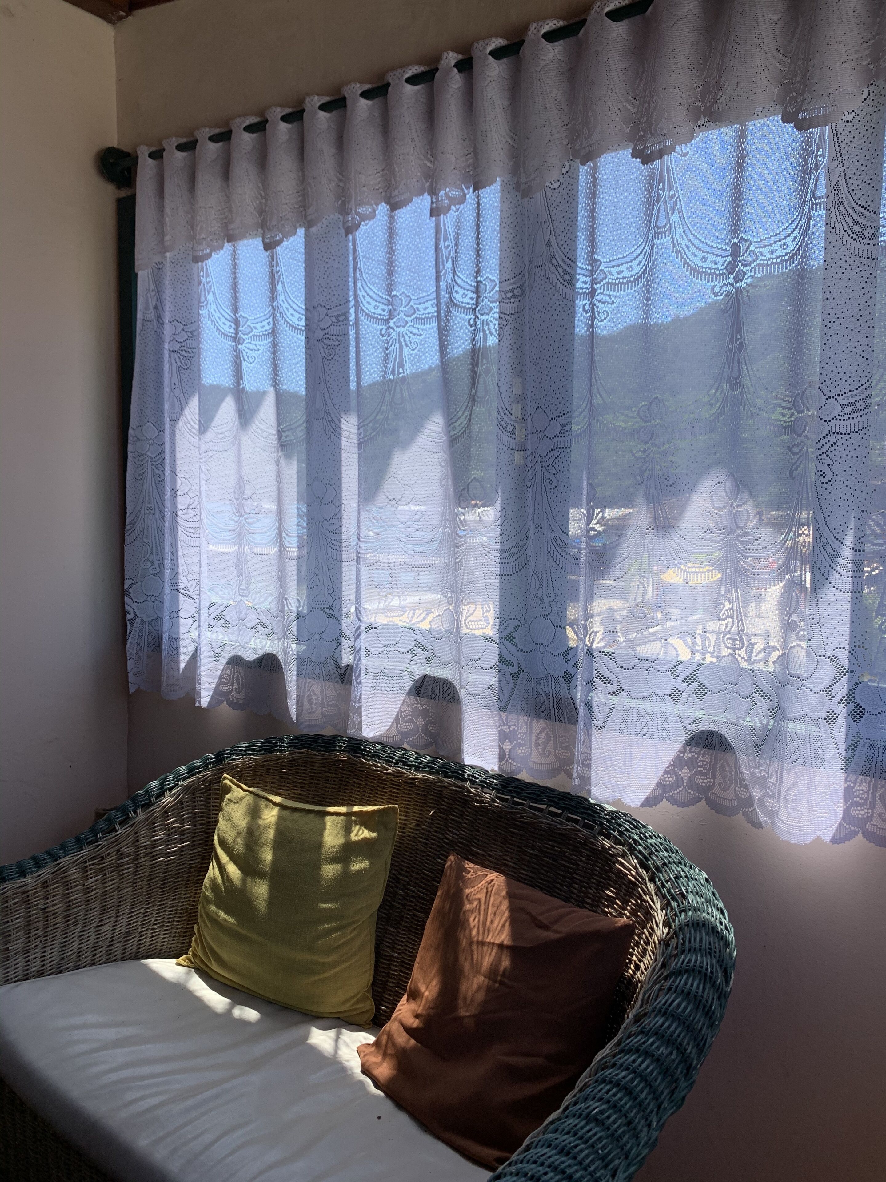 Um sofá de vime com almofadas, uma janela com cortinas de renda e vista para um edifício.