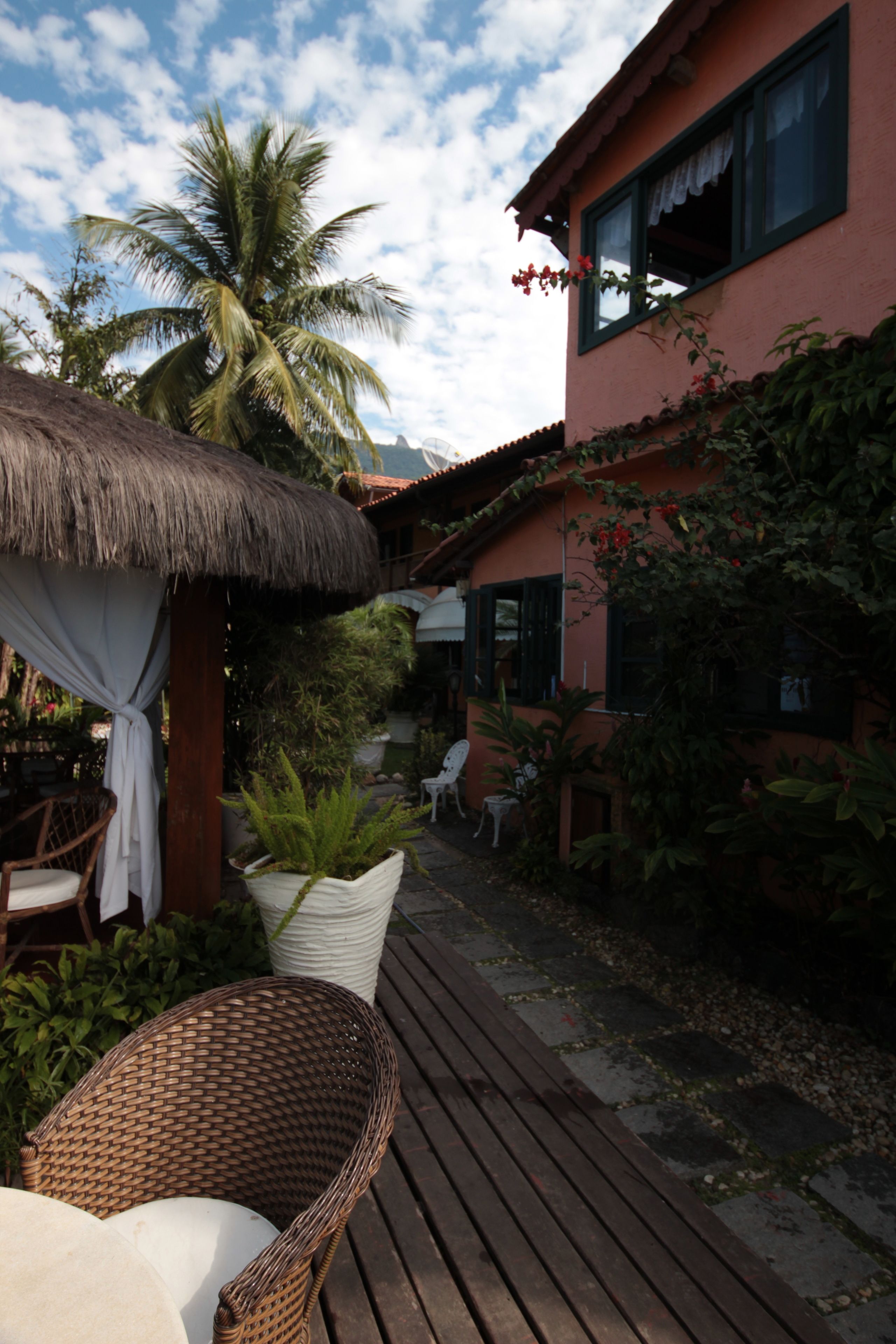 A wooden deck with wicker chairs, a thatched-roof structure, and a pink building with windows and plants.