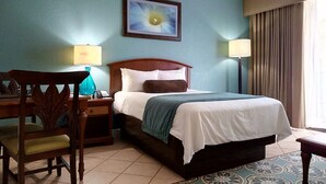 A hotel room with a bed, a desk, a chair, two lamps, a painting on the wall, and a window with curtains.