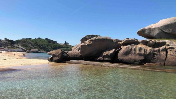 Uma praia com águas calmas e transparentes, pedras grandes e uma faixa de areia.