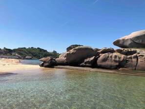 A beach with clear shallow waters, large rocks, and a sandy shore.