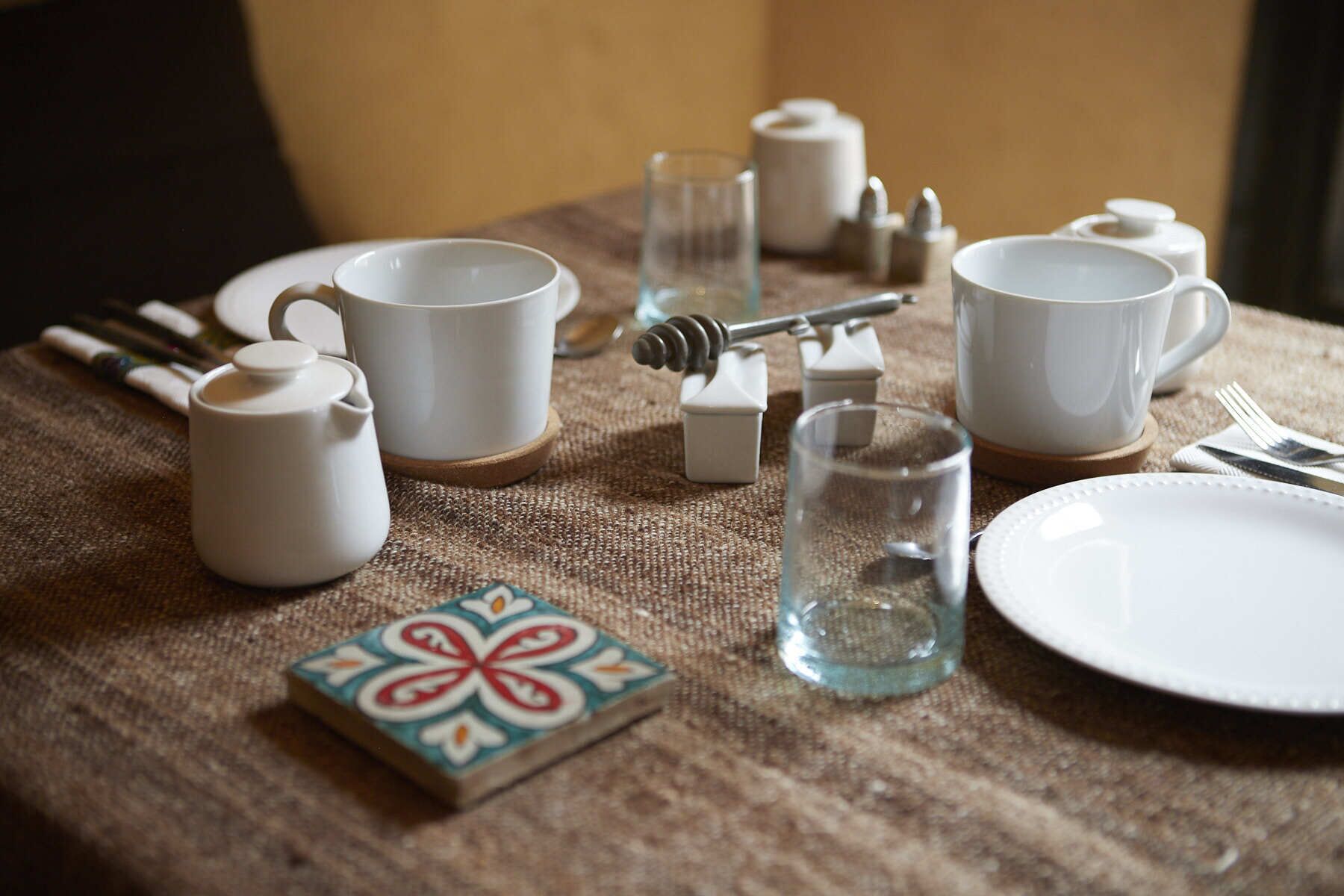 A table set with white ceramic cups, a teapot, a glass, and a plate, along with a honey dipper and a small decorative tile.