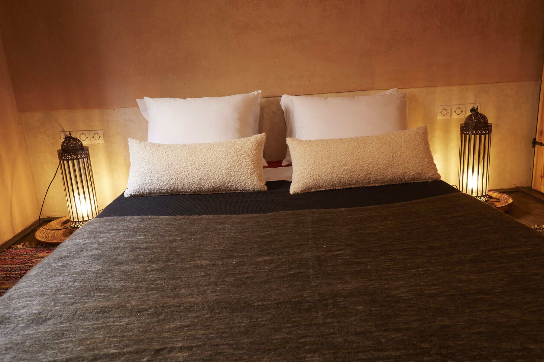 Una cama con dos almohadas y una manta oscura, con dos lámparas fijadas a la pared.