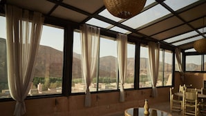 A room with large windows, a woven chair, and a glass-top table. - Kasbah Flilou (Ait Sedrate Jbel El Soufla)