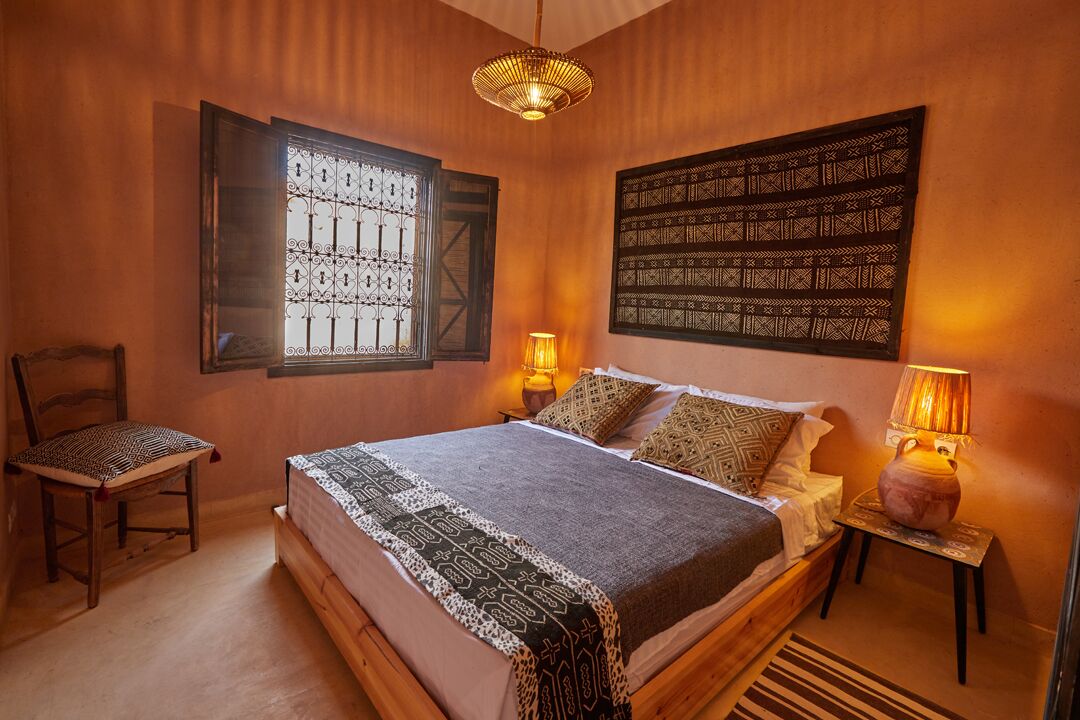 A cozy bedroom with a wooden bed, a chair, a window with decorative iron bars, and a wall hanging.