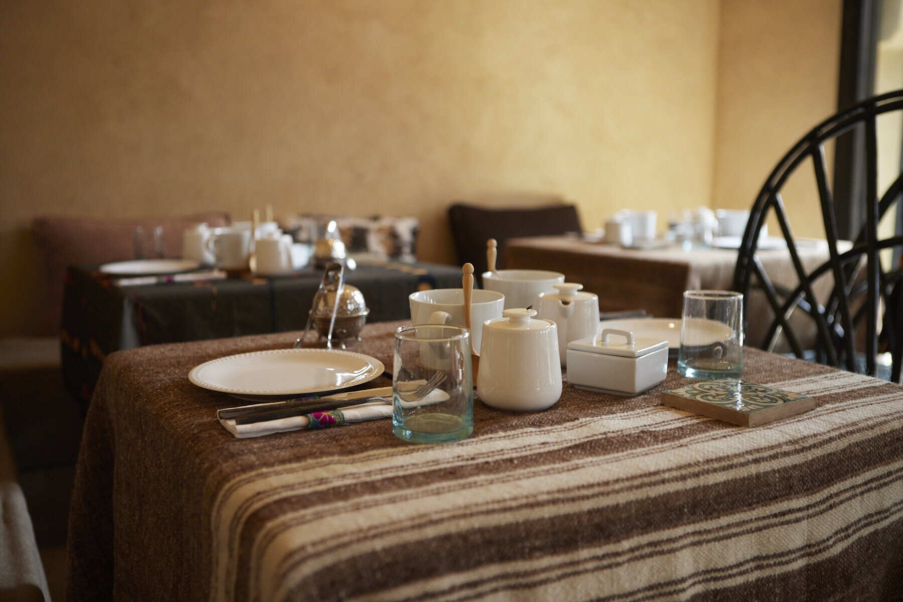 Une table à manger dressée avec de la vaisselle blanche, des verres et une théière, recouverte d’une nappe à rayures, et une chaise placée au premier plan.