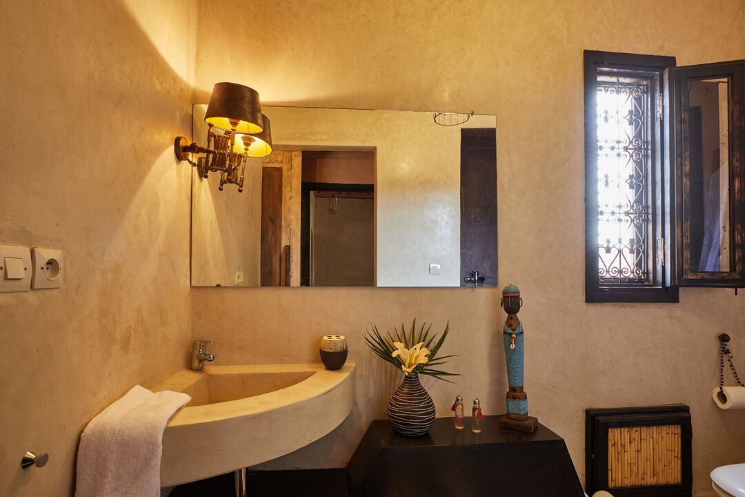 Un baño con un lavamanos curvo, un espejo grande, una lámpara de pared, un jarrón decorativo y un inodoro con panel de bambú.