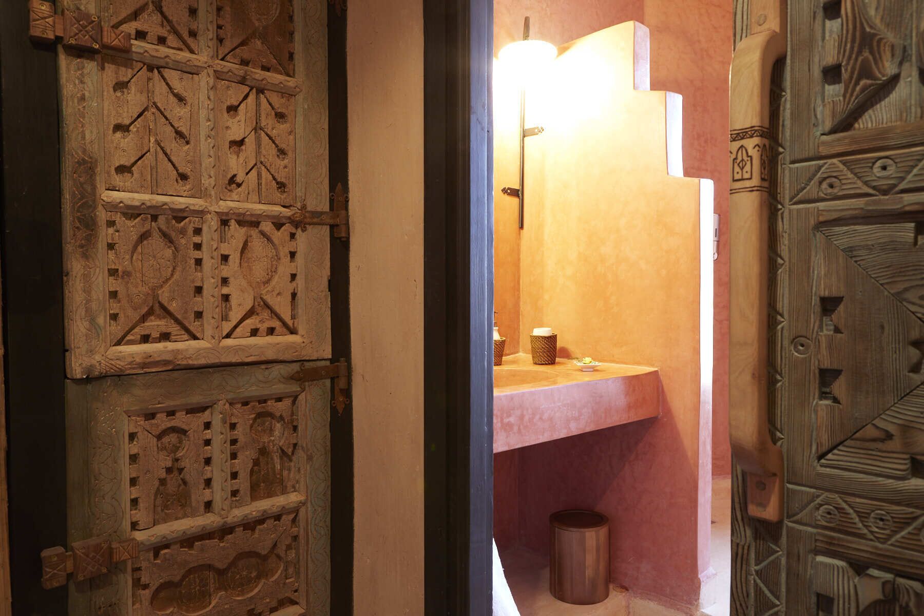 A wooden door with intricate carvings, a pink sink with a mirror above it, and a trash can in a bathroom.