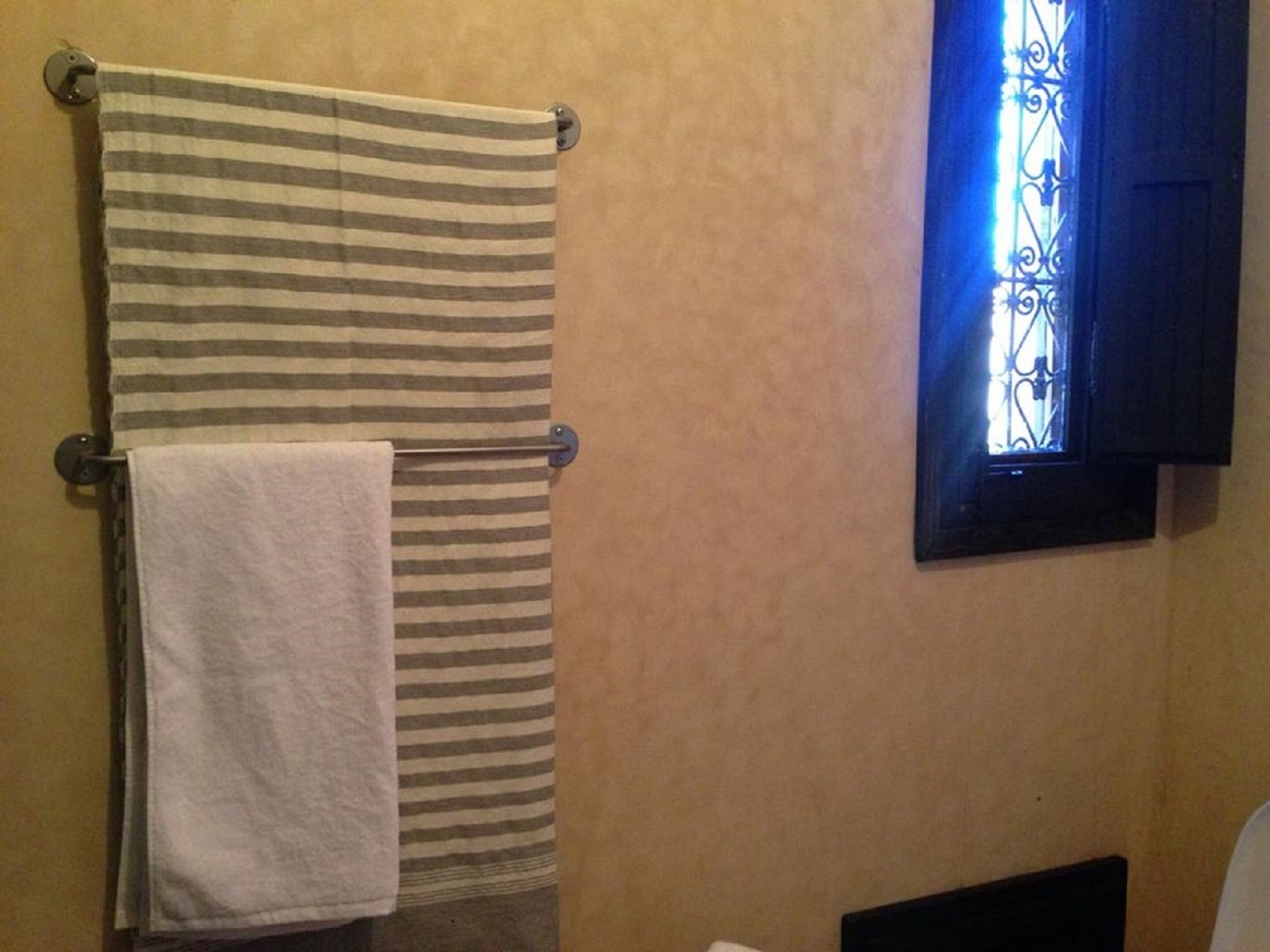 Een badkamer met een gestreept handdoek aan een handdoekhouder, een effen witte handdoek en een raam met decoratieve ijzeren tralies.