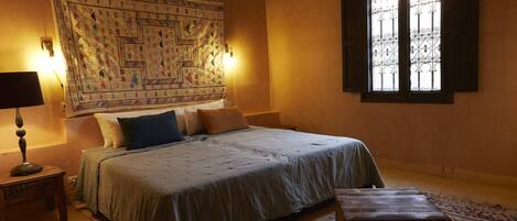 Un dormitorio con una cama grande, dos lámparas de noche, un tapiz en la pared y un ventanal con rejas.