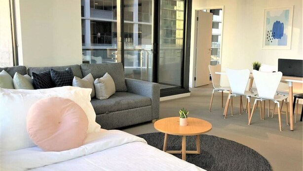 Una habitación de hotel moderna con cama, sofá gris, una pequeña mesa redonda con una planta y un comedor con sillas y mesa blancos.