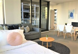 A modern hotel room with a bed, a grey sofa, a small round table with a plant, and a dining area with white chairs and a table. - Central Location & WiFi (Melbourne)