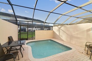 A glass-enclosed swimming pool area with a table and chairs, a tiled floor, and a view of a building in the background.