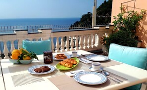 A table set for breakfast with croissants, a bowl of fruit, and a jar of jam, overlooking a scenic view of the sea.