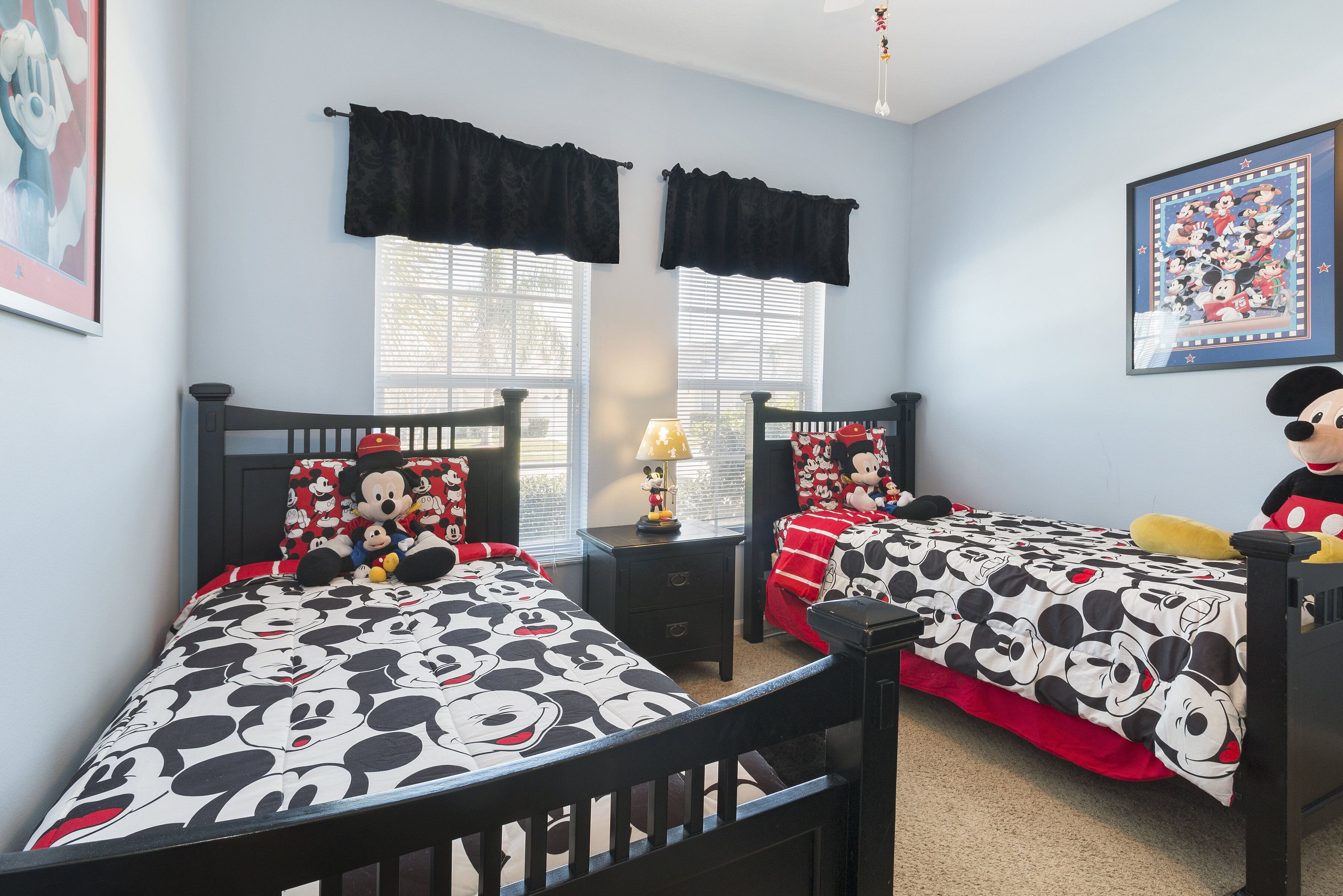 A bedroom with two beds, each adorned with Mickey Mouse-themed bedding. There are two windows with black valances, a nightstand with a lamp, and a framed picture on the wall.