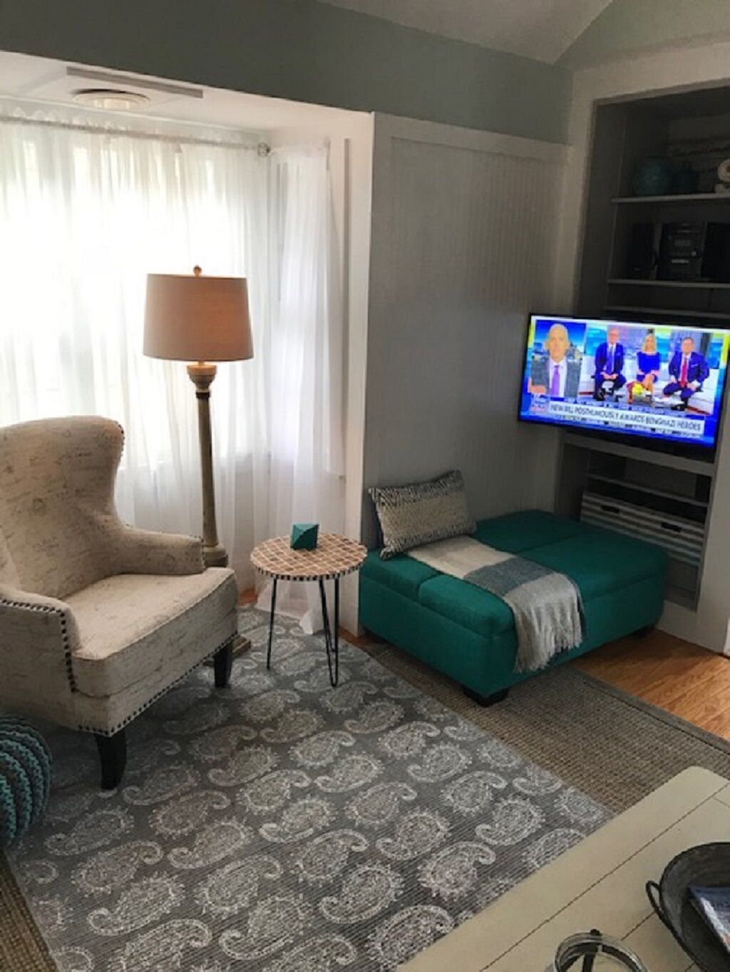 A living room with a beige armchair, a small round table, a teal sofa, and a television displaying a news program.