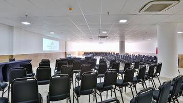 Un salón de conferencias con filas de sillas negras, un atril y una pantalla de proyector mostrando el nombre del hotel.