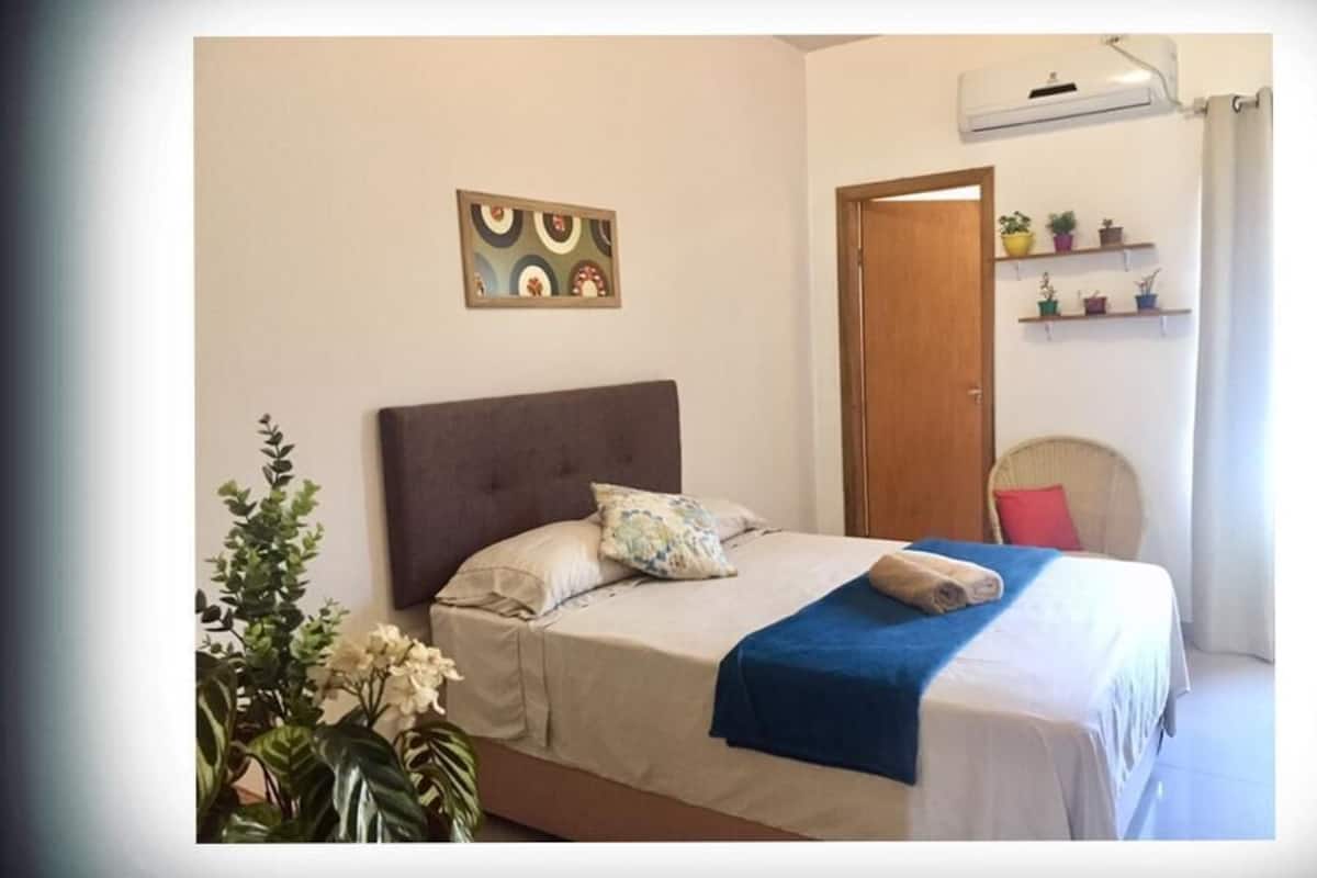 A bedroom with a bed, a headboard, a wall-mounted shelf with potted plants, a door, and a window with curtains.