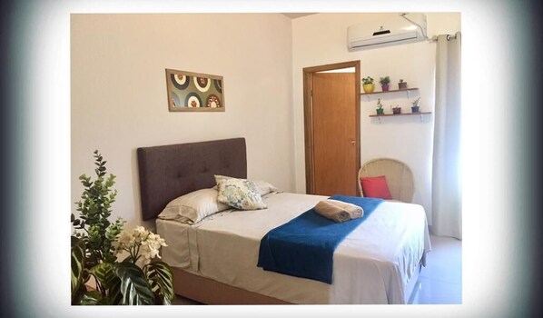 A bedroom with a bed, a headboard, a wall-mounted shelf with potted plants, a door, and a window with curtains.