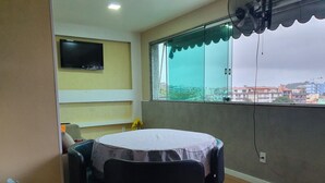 Ein Zimmer mit einem runden Tisch, der mit einer Tischdecke bedeckt ist, zwei schwarzen Stühlen, einem an der Wand befestigten Fernseher und einem Fenster mit Blick auf Gebäude.
