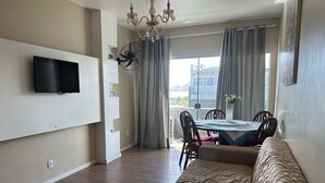 Una sala de estar con un televisor de pantalla plana montado en la pared, un comedor con mesa y sillas, y un ventanal con cortinas.