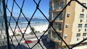 Vista a través de una cerca de alambre que da a una playa, una carretera con un coche rojo y un edificio de varios pisos con balcones.