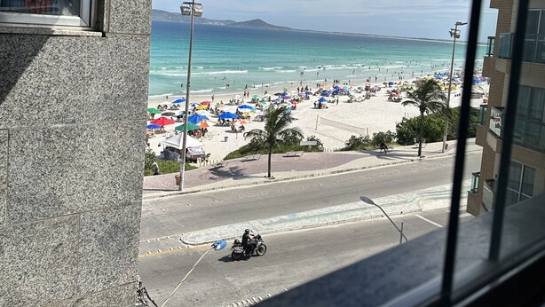 Vista de una playa con sombrillas y gente, una carretera con una motocicleta y edificios al fondo.
