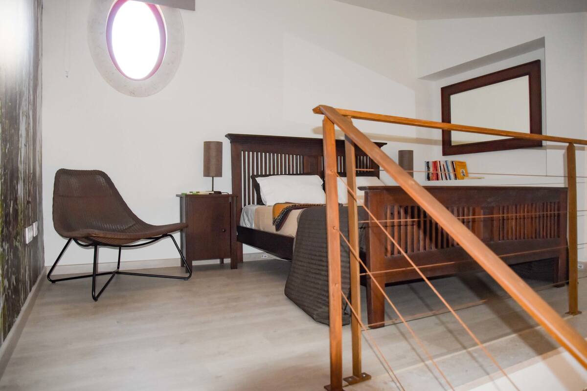 A bedroom with a bed, a chair, a nightstand, a mirror, and a staircase.