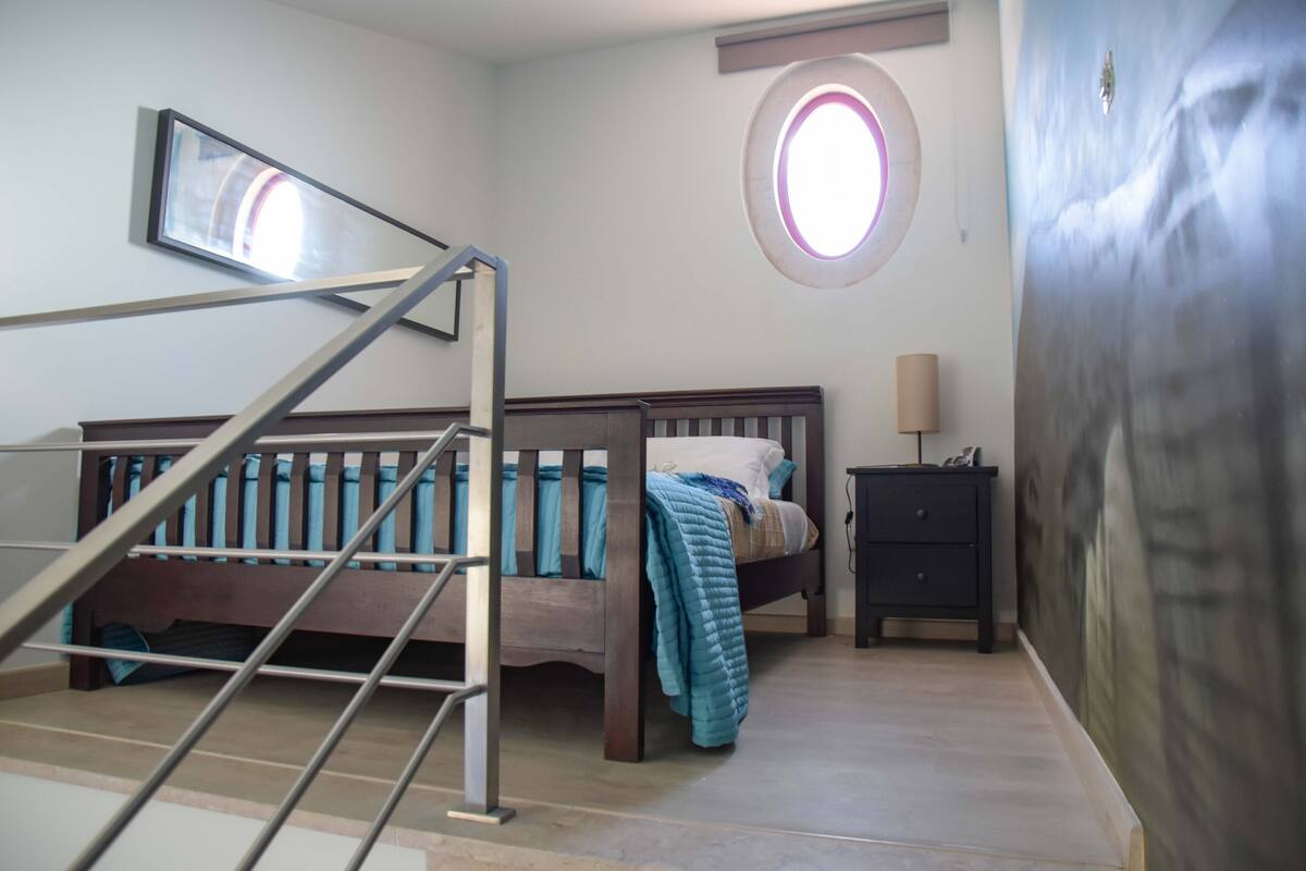 A bedroom with a wooden bed, a nightstand, a lamp, a window with a view, and a staircase.