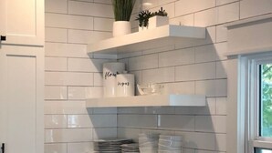 Une cuisine avec des murs recouverts de carreaux blancs, des meubles de cuisine blancs et un plan de travail noir. On y trouve des étagères blanches où sont rangés des ustensiles de cuisine, et une fenêtre sur la droite.