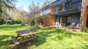 A backyard with a wooden picnic table, a brick house with a balcony, and a garden with flowering trees.