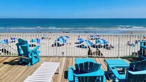 Blick auf einen Strand mit einer Holzterrasse, blauen Adirondack-Stühlen und Sonnenschirmen.