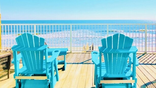 Eine Holzterrasse mit zwei blauen Adirondack-Stühlen, die auf einen Strand ausgerichtet sind.