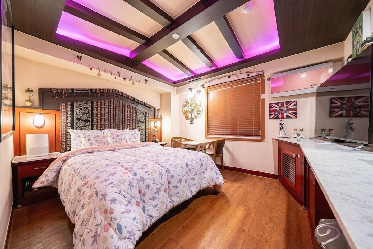 Une chambre avec un grand lit, un plancher en bois, un bureau et une fenêtre avec des stores.