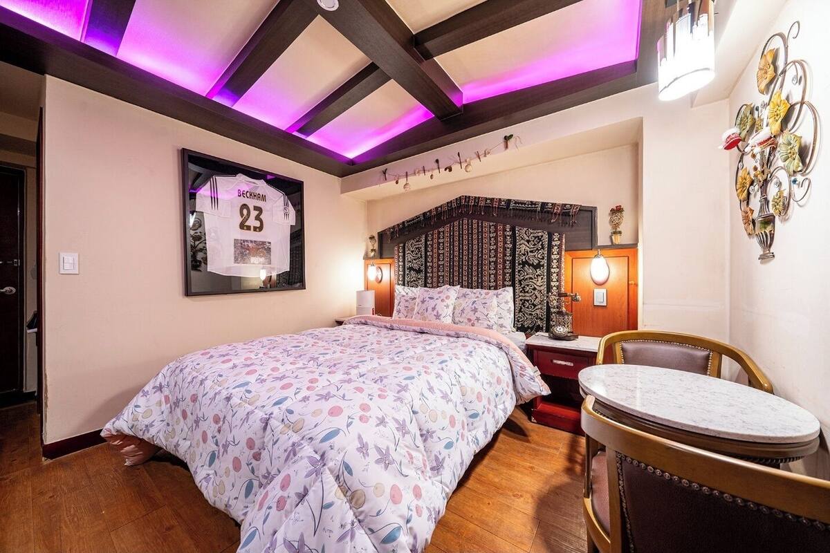 A bedroom with a bed, a chair, a table, and a wall with a framed jersey.
