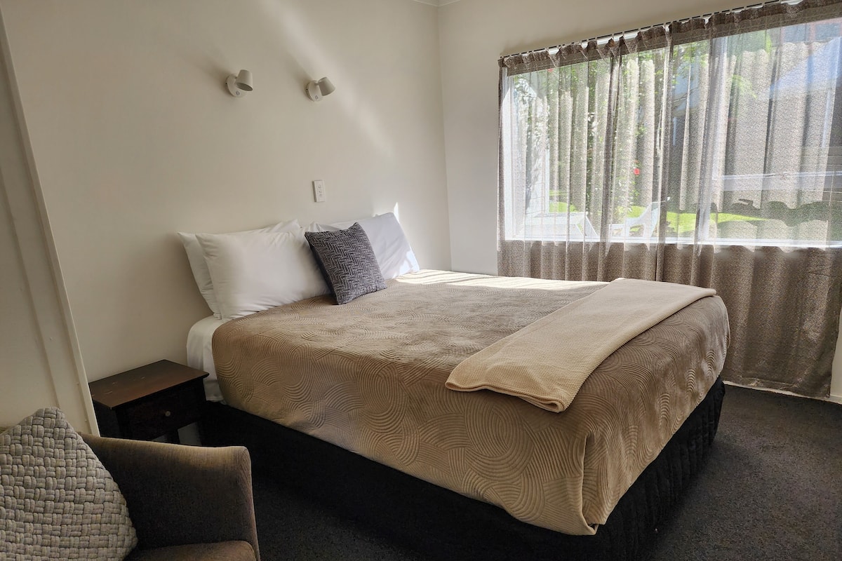 A neatly made bed with a patterned bedspread, a bedside table, a chair, and a window with sheer curtains.