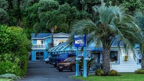 Um motel com fachada azul, estacionamento com vários carros e vegetação exuberante ao redor.