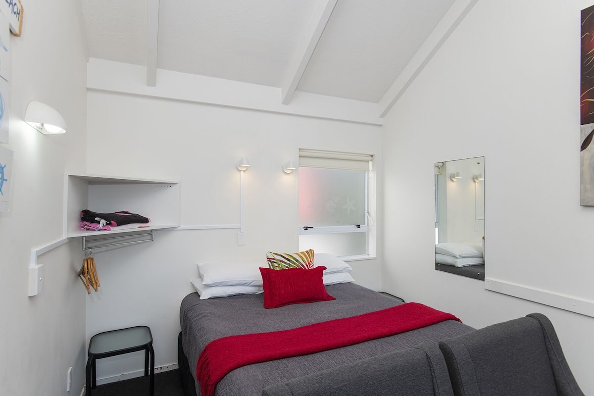 A bedroom with a bed, a bedside table, a chair, a shelf with items, a window, and a mirror.