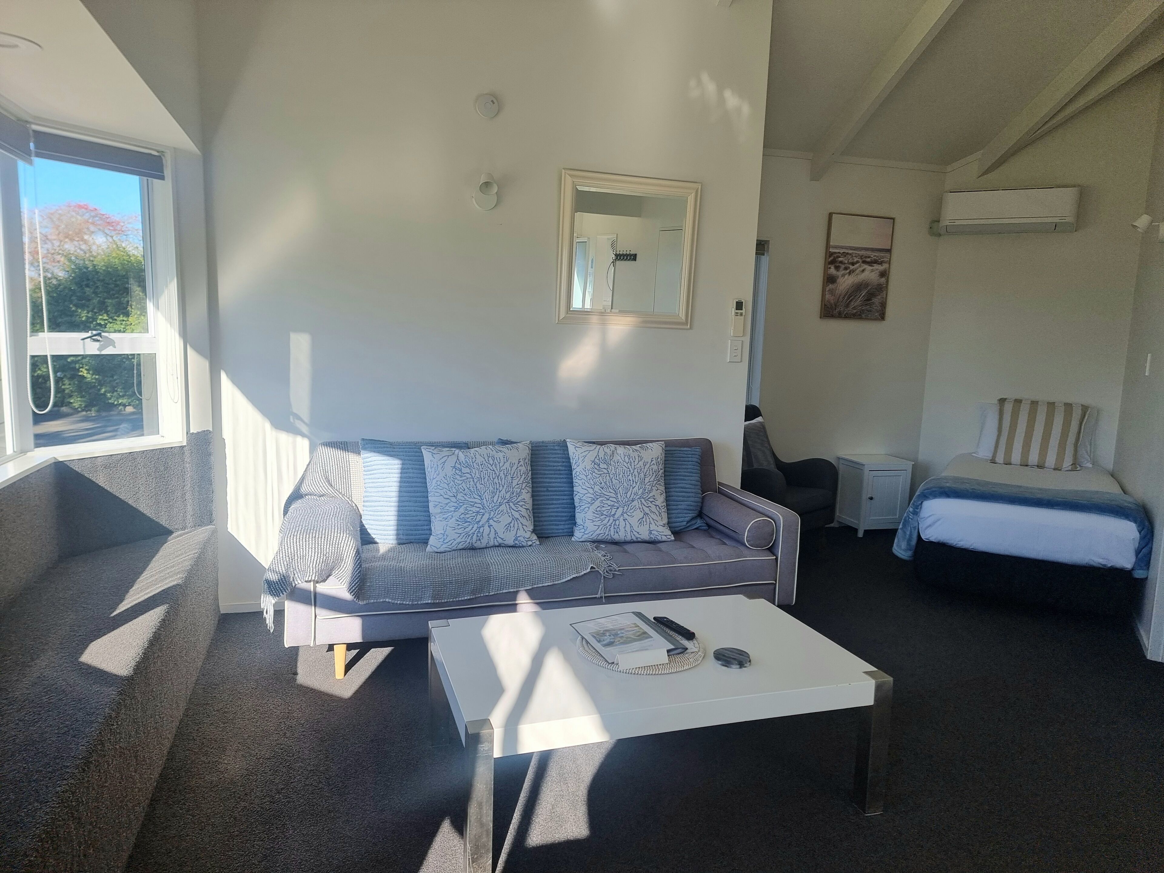 A room with a sofa, a small table, a bed, a mirror, and an air conditioning unit.