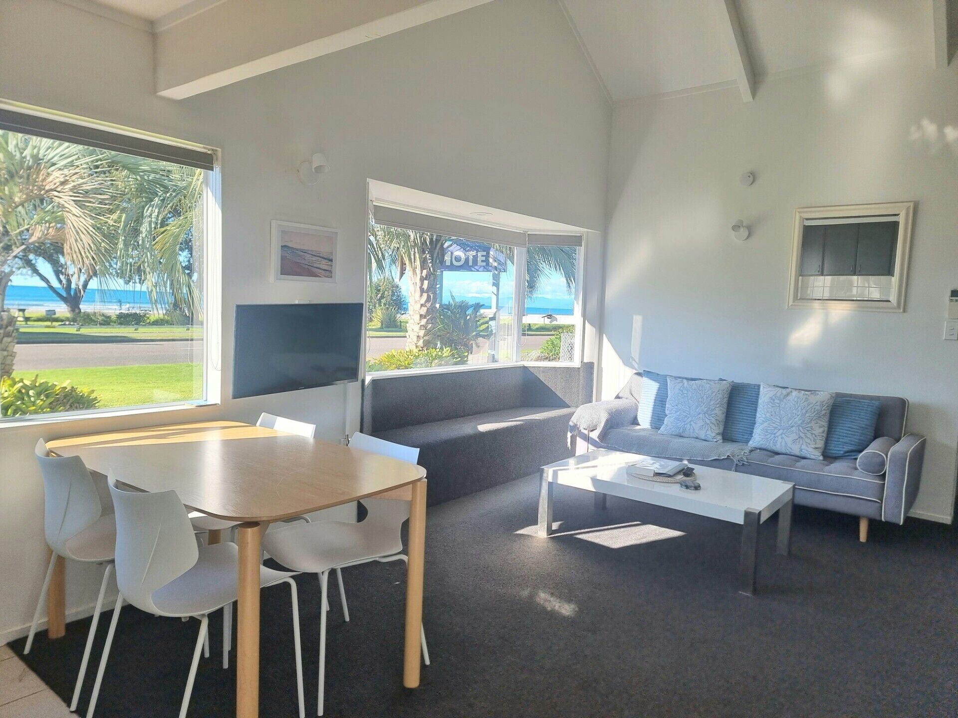 Una sala moderna con comedor, un sofá y una mesa de centro. Hay un ventanal con vistas a la playa y palmeras.
