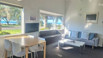 Una sala moderna con comedor, un sofá y una mesa de centro. Hay un ventanal con vistas a la playa y palmeras.
