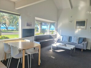 A modern living room with a dining area, a sofa, and a coffee table. There is a large window with a view of the beach and palm trees. - Ohope Beach Motel (Ohope)