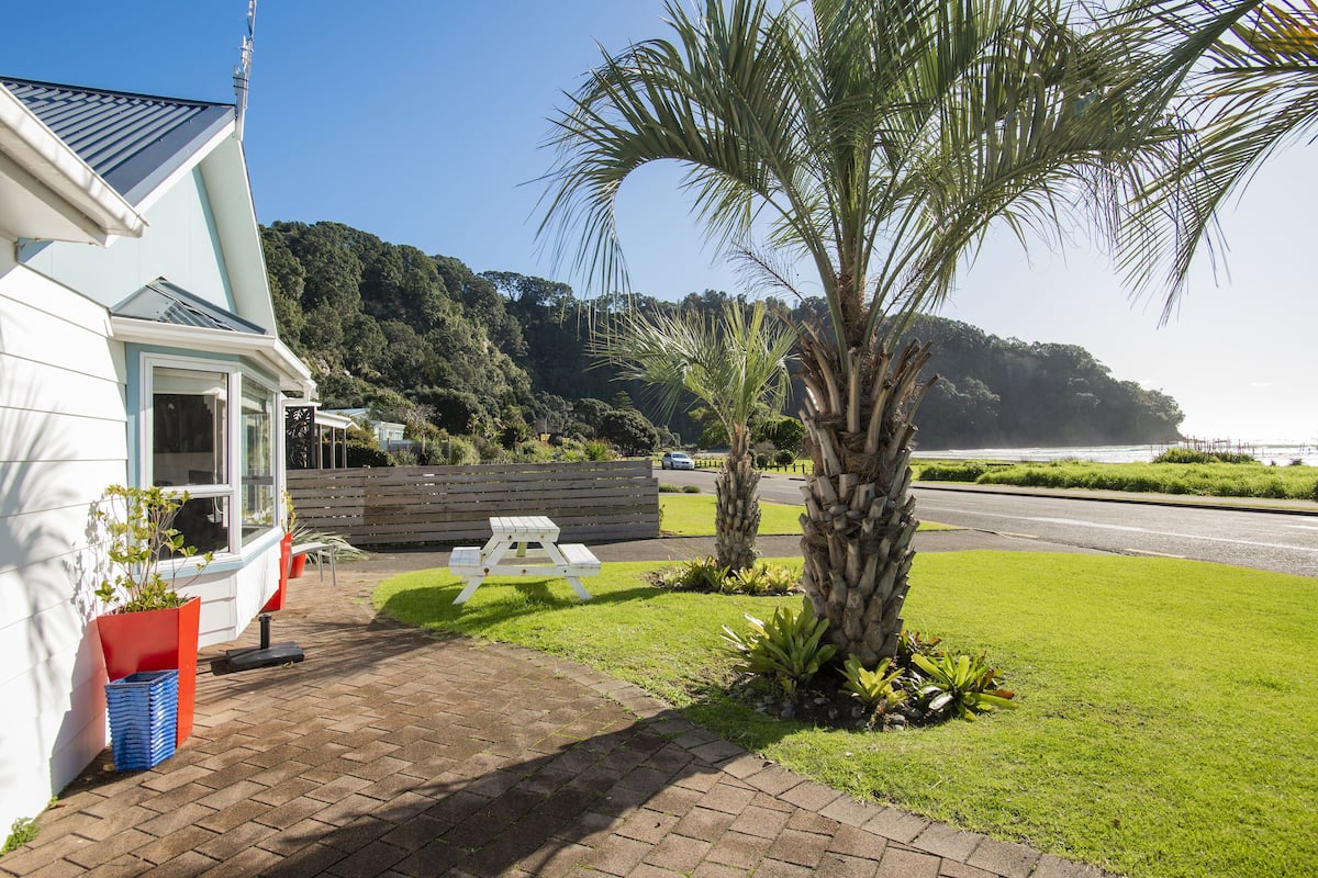 A coastal property with a white building, a paved patio, a white picnic table, and a palm tree.