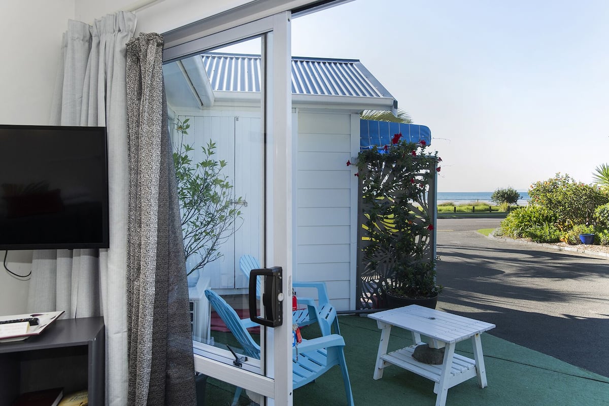 A room with a TV, a small table, and chairs, a sliding glass door leading to a balcony with a view of the sea.