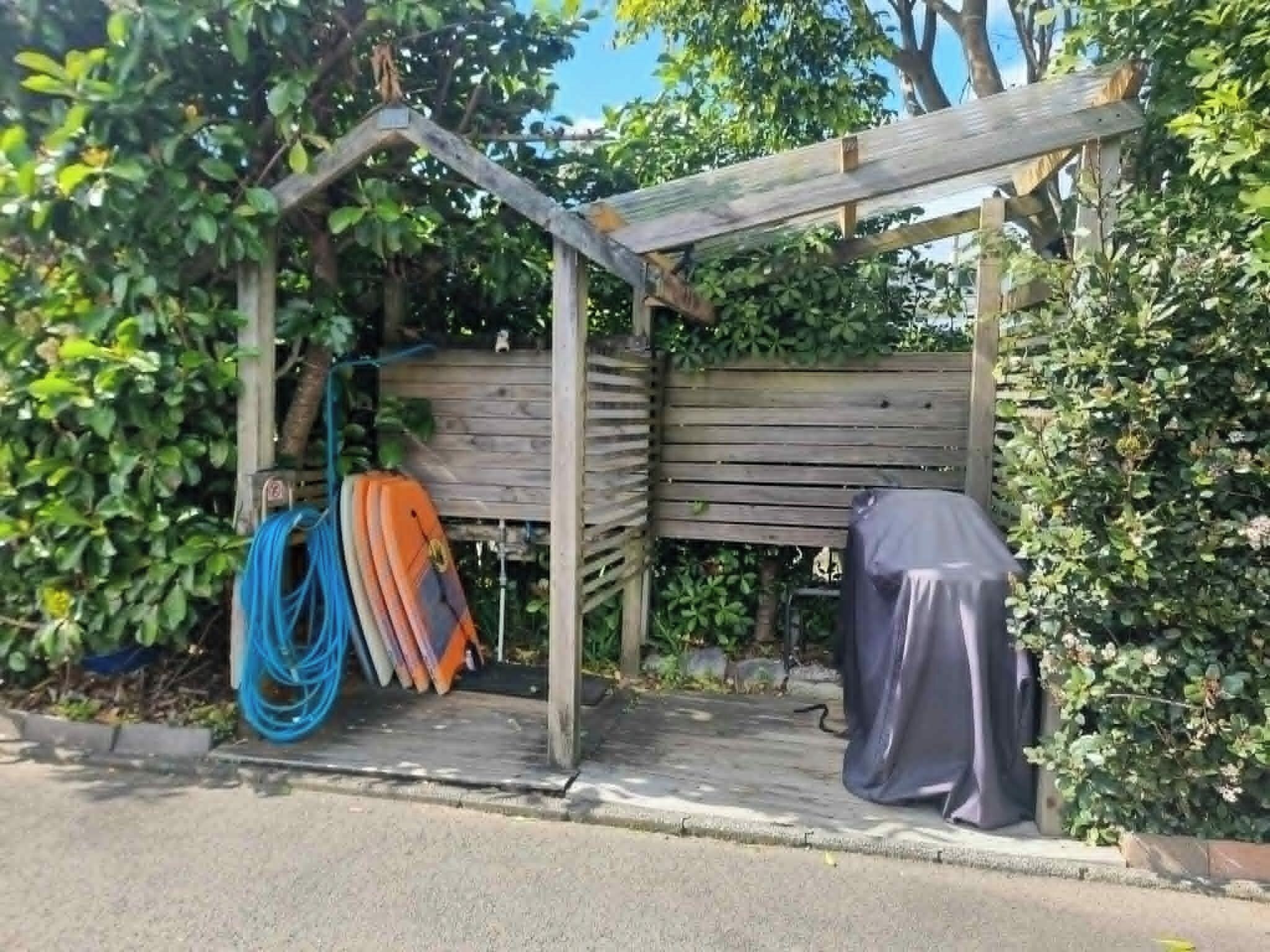 Una estructura de madera con un banco, rodeada de vegetación, y se observan tablas de surf y una manguera.