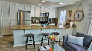 A kitchen with white cabinets, a stainless steel refrigerator, a microwave, and a granite countertop. There is a dining area with a blue table and a gray sofa. - Luna Azul, only steps to Mission Beach! Free Wireless Internet / NO SMOKING (San Diego)