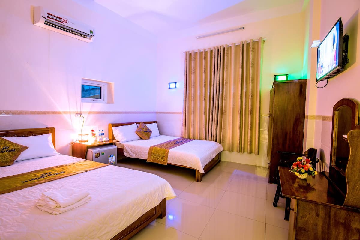 A hotel room with two beds, a TV, a wardrobe, and a desk with a lamp and flowers.