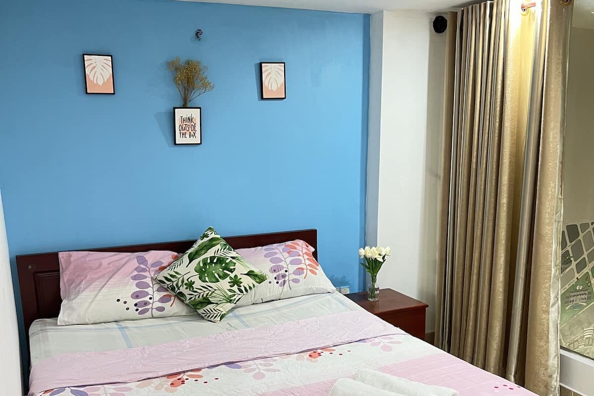 A neatly made bed with floral bedding, a bedside table with a vase of flowers, and a blue wall with framed artwork.